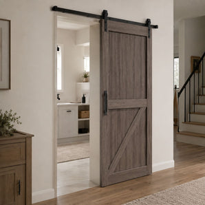 A rustic gray-brown wooden sliding barn door with a black metal rail and hardware, shown installed in a bright, modern home entryway.