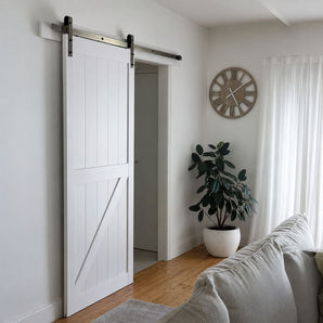 A white barn-style sliding door with dark metal hardware installed in a bright room with light wood floors, a potted plant, a wall clock, and part of a sofa visible.