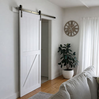 A white barn-style sliding door with dark metal hardware installed in a bright room with light wood floors, a potted plant, a wall clock, and part of a sofa visible.